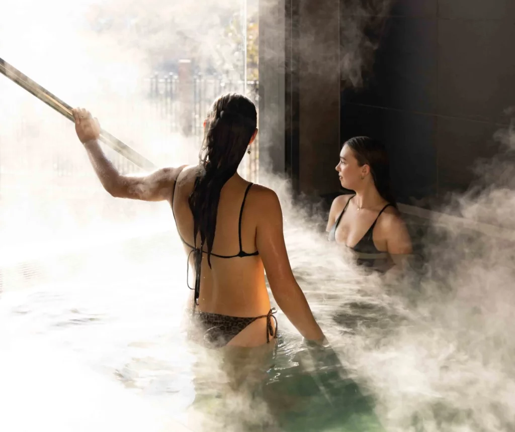 Two girls standing in a steaming private geothermal pool at Wai Ariki, holding onto a metal railing as steam rises around them.