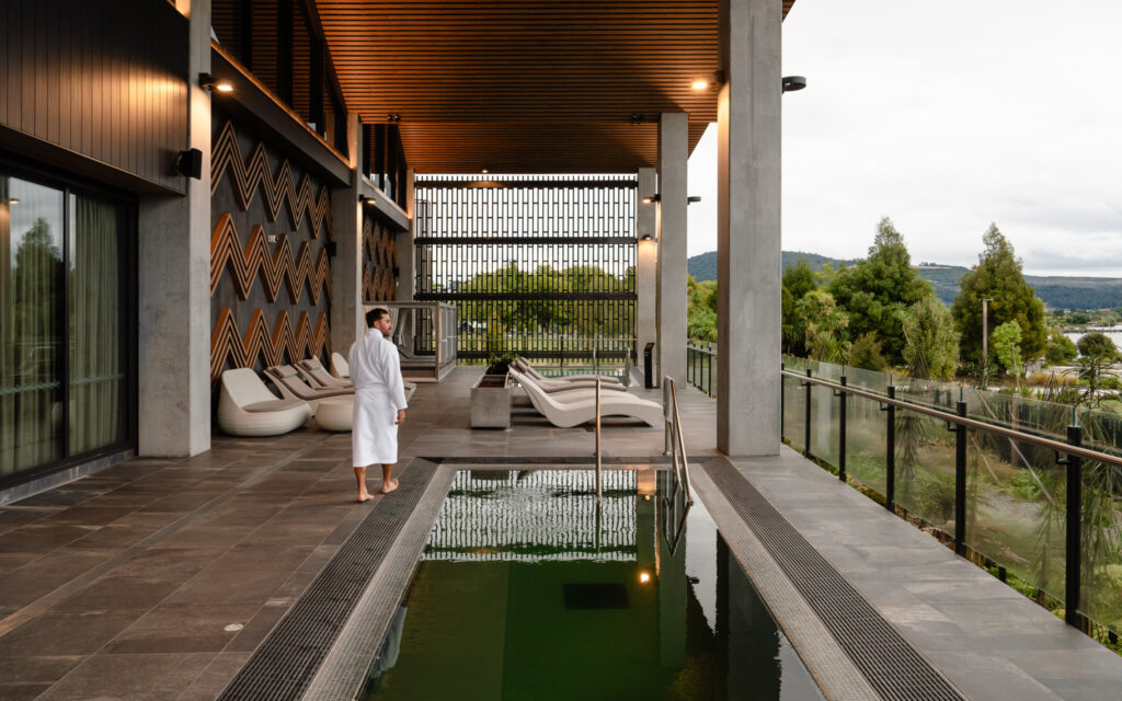 Guest in a robe walking along Wai Ariki's Sanctuary concourse, beside a goethermal pool with views over the surrounding grounds and Lake Rotorua.