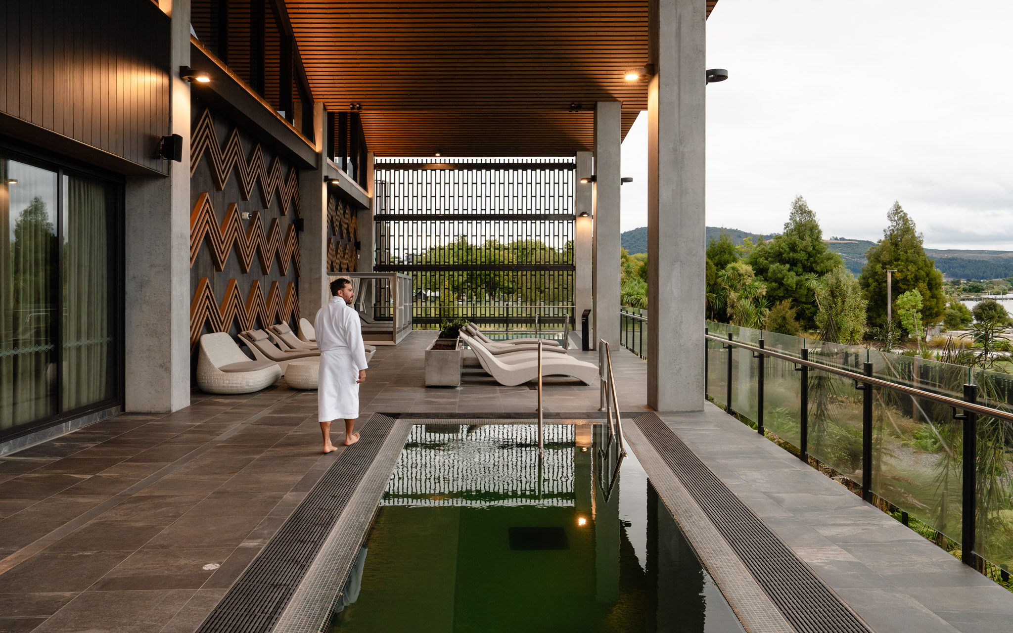 Guest in a robe walking along Wai Ariki's Sanctuary concourse, beside a goethermal pool with views over the surrounding grounds and Lake Rotorua.