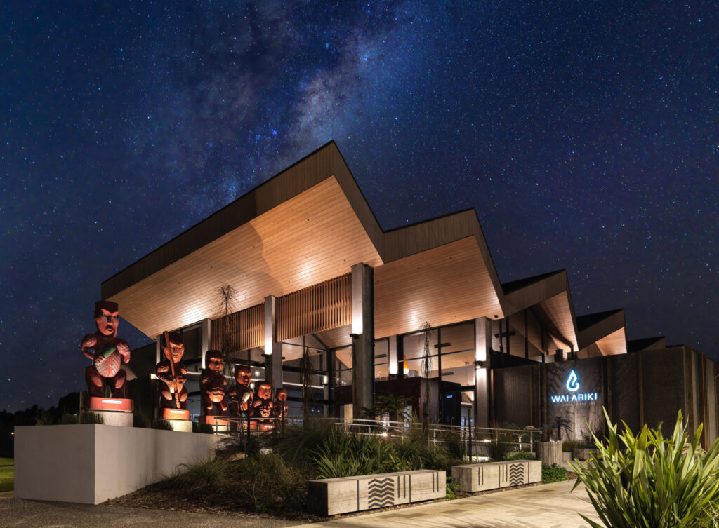 Wai Ariki Hot Springs and Spa entrance at night, with carved pouwhenua (carved wooden figures) lit beneath the building's wing-like wooden canopy, set against a clear night sky showing the Milky Way.