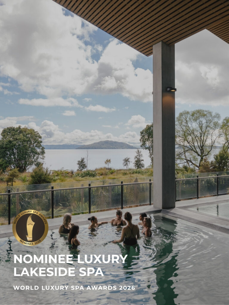 A group of people relax together in an outdoor thermal pool at Wai Ariki Hot Springs and Spa, with views across to Mokoia Island on Lake Rotorua. Gold badge reads "Nominee Luxury Lakeside Spa, World Luxury Spa Awards 2026."