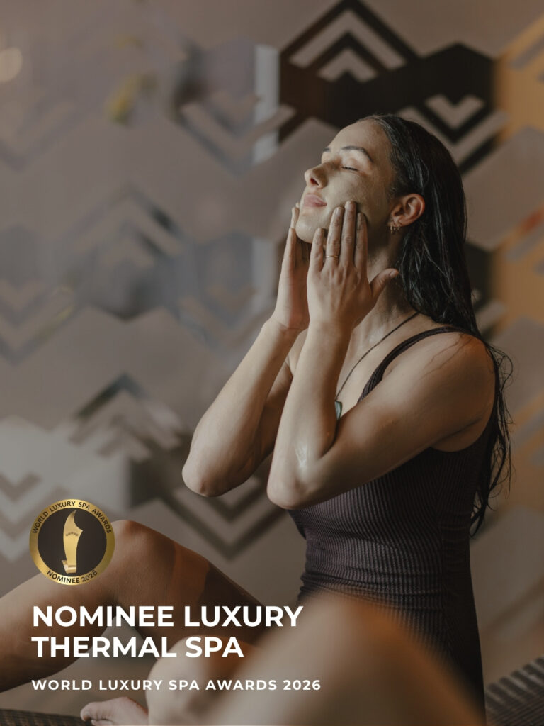 A woman with a clay mud mineral mask applies it to her face with both hands, eyes closed, set against a patterned wall backdrop. Gold badge reads "Nominee Luxury Thermal Spa, World Luxury Spa Awards 2026."