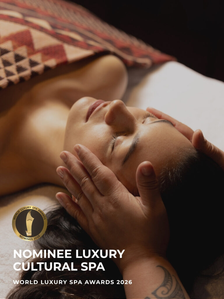 A woman lies on a treatment table on a woven Māori-patterned pillow while a therapist's hands, one with a tā moko (Māori tattoo), gently cradle her face. Gold badge reads "Nominee Luxury Cultural Spa, World Luxury Spa Awards 2026."