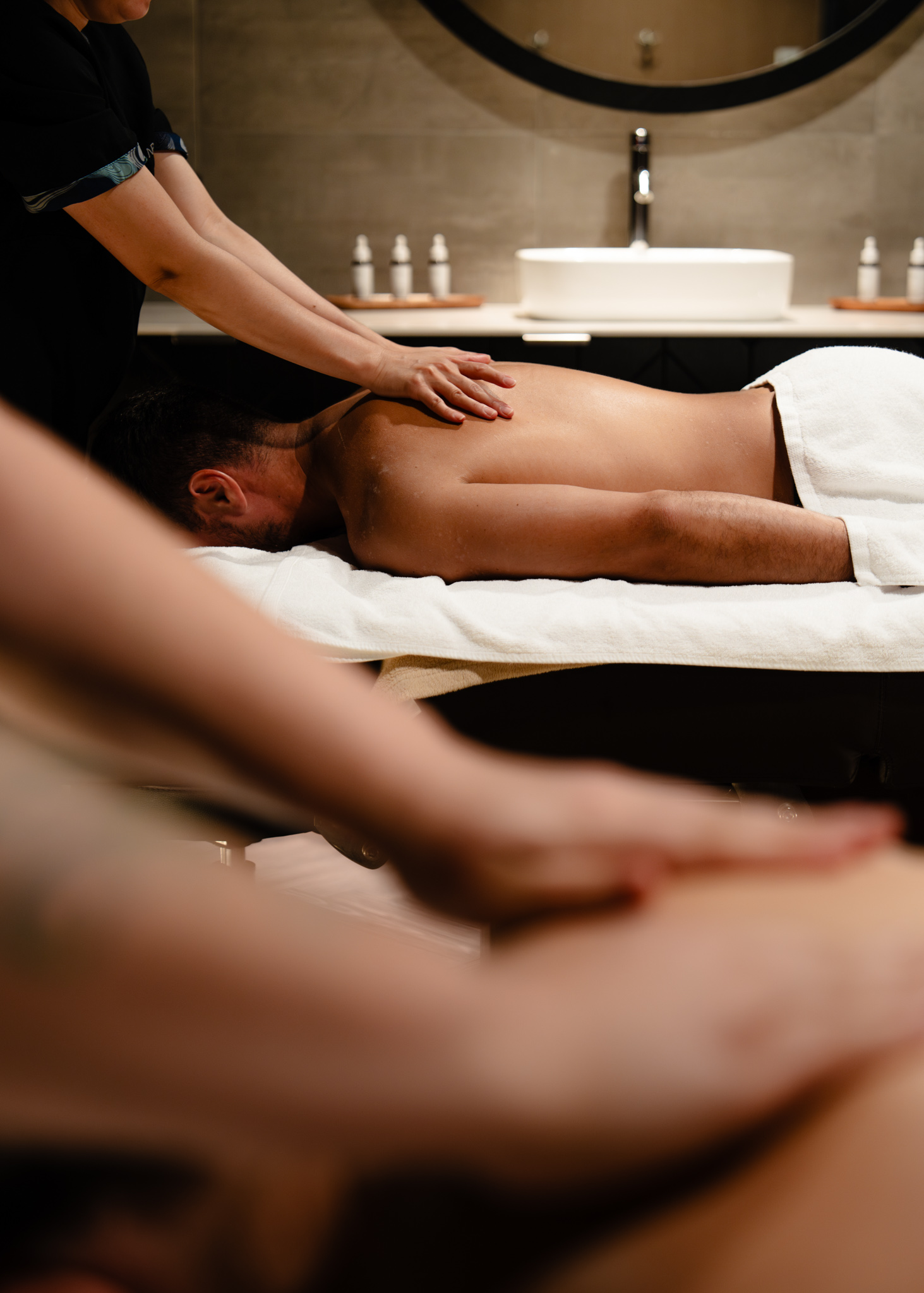 Guest receiving a back massage from a therapist in a Wai Ariki treatment room, with amenities and a vanity basin in the background.