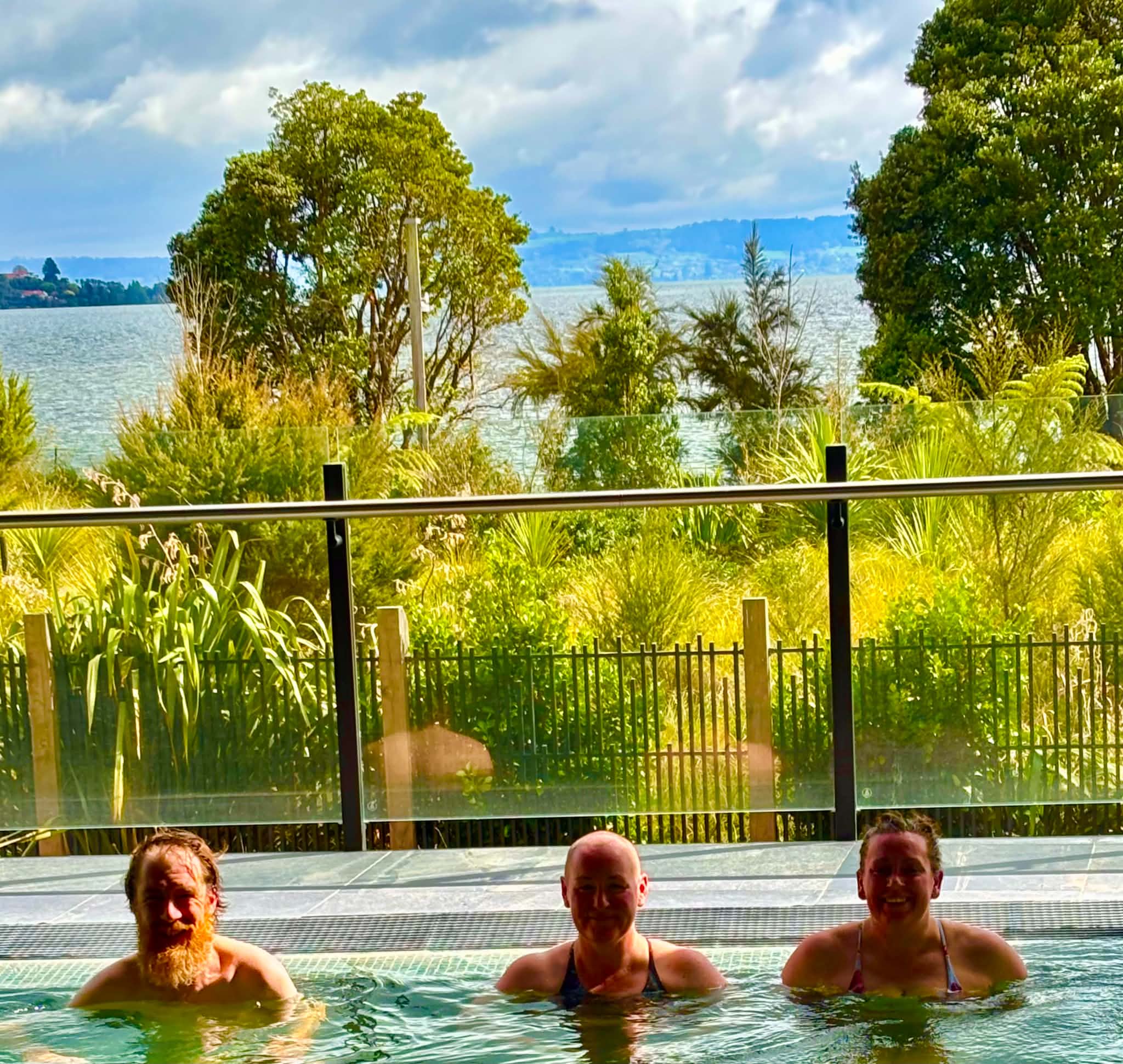 Guests soaking in a geothermal pool beneath open skies, with lake views and contemporary architecture at Wai Ariki Hot Springs and Spa in Rotorua.