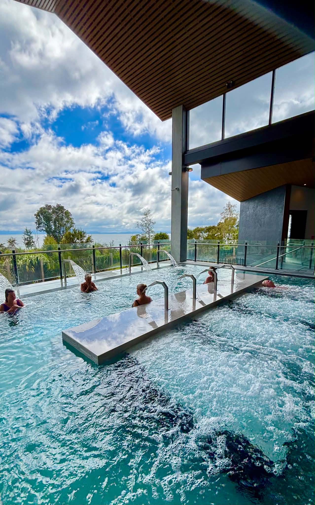 Three guests relaxing in a geothermal pool with views of Lake Rotorua and native gardens at Wai Ariki Hot Springs and Spa.